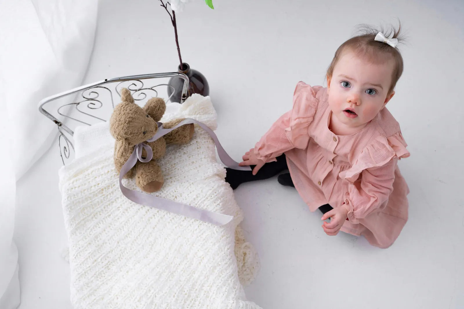 A toddler in a pink dress sitting beside a teddy bear on a knitted blanket